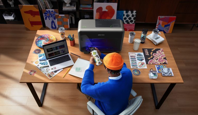 eufyMake E1 centered on a workbench surrounded by colorful artwork, decorated drinkware and small personalized projects.